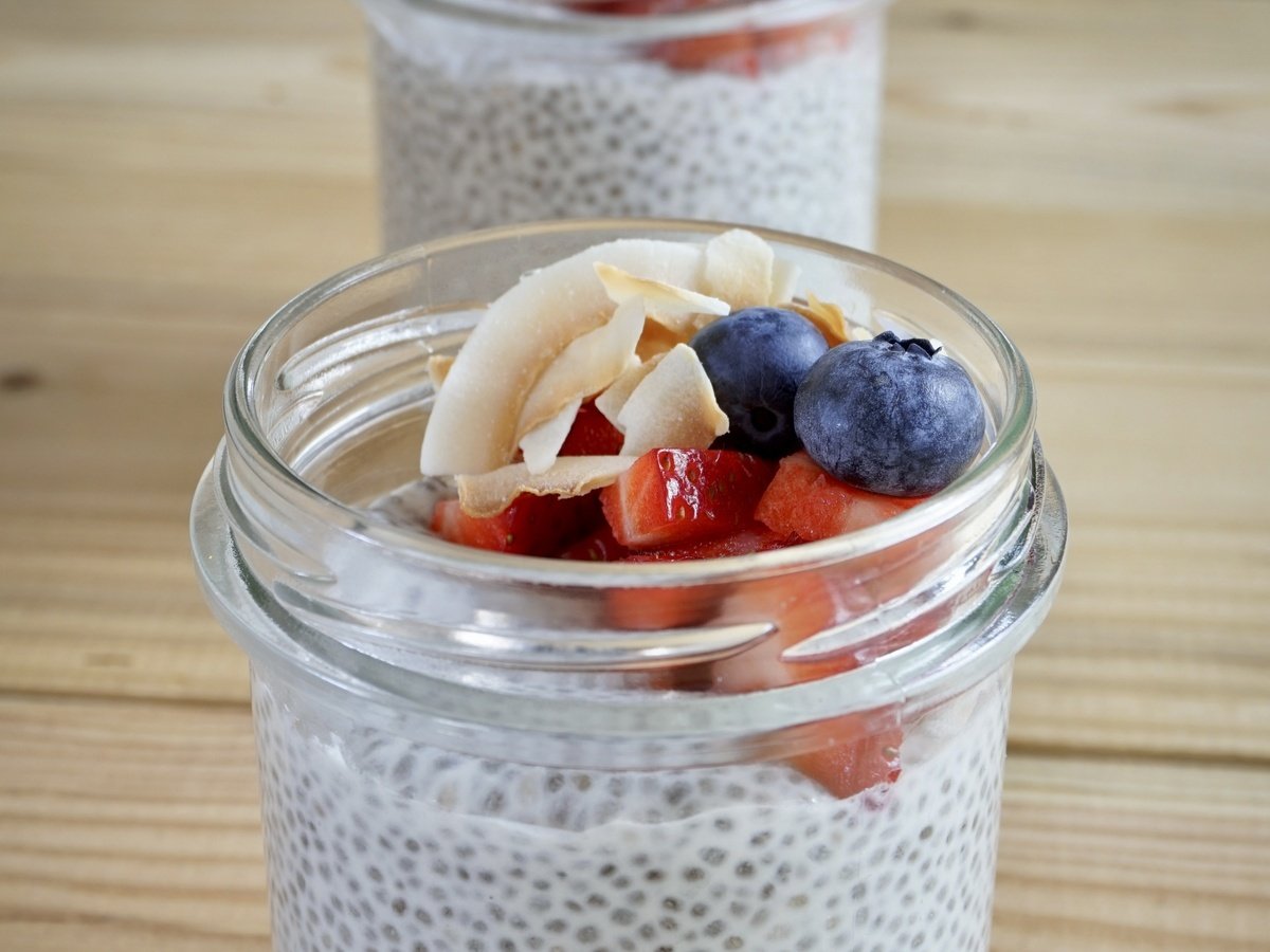 Balang kaca puding biji chia berkrim ditaburi beri biru, strawberi segar dan kepingan kelapa di atas meja kayu